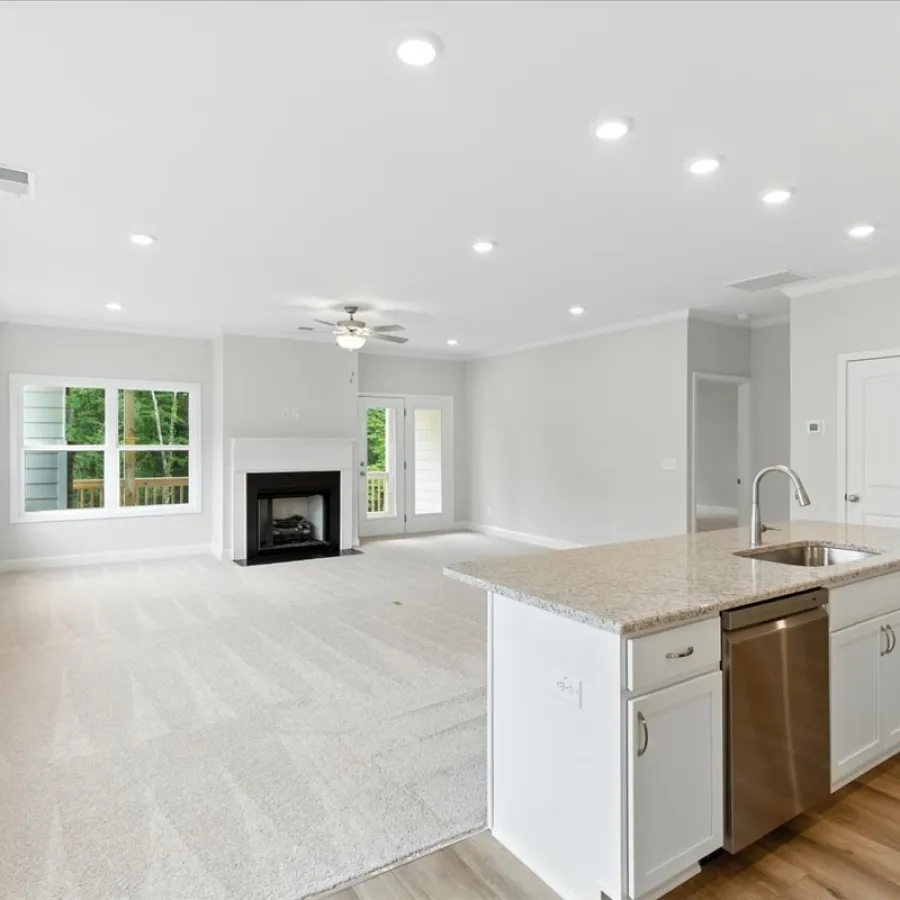 Bright open living room with white walls, large windows, carpeted floor, and kitchen island with sink and dishwasher.