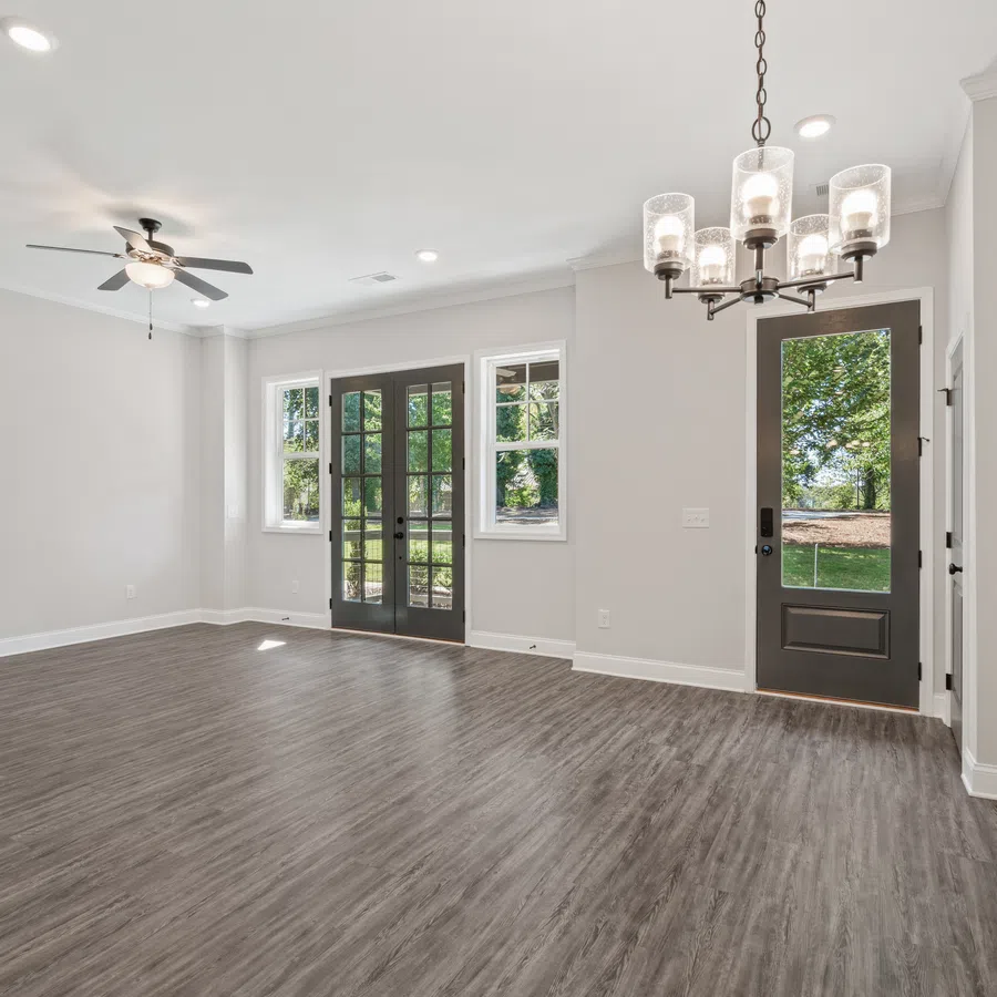 Empty modern room with gray wood flooring, ceiling fan, chandelier, and glass doors leading outside