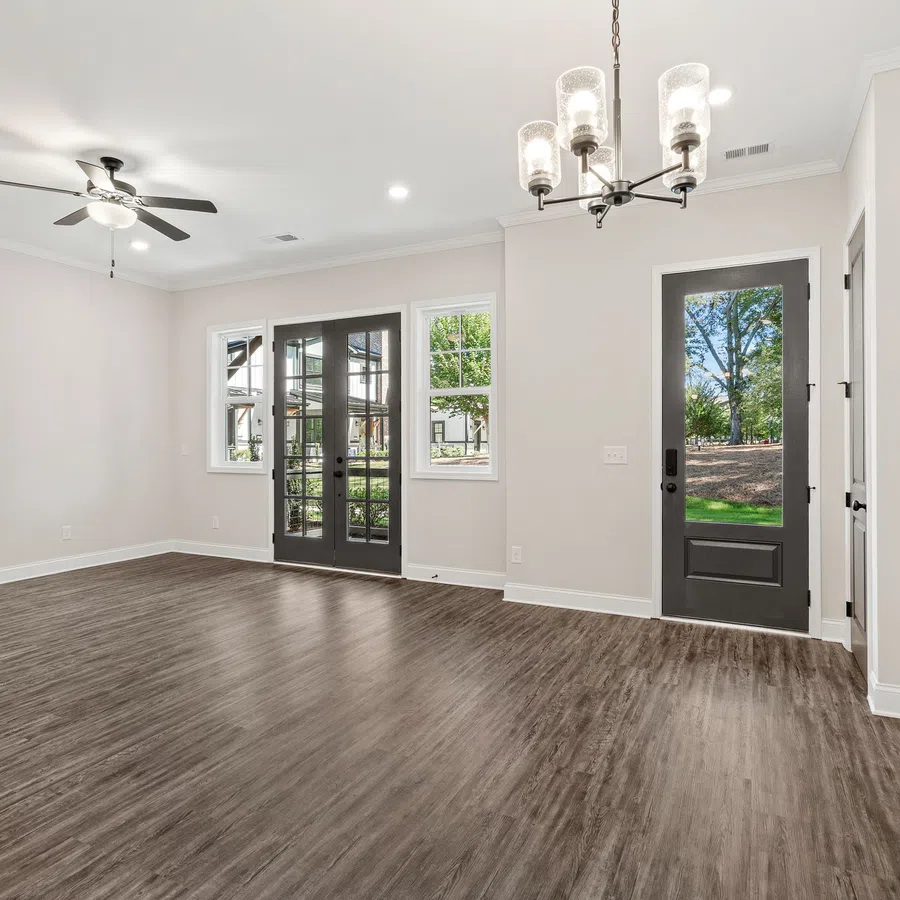 Empty modern room with wood flooring, ceiling fan, chandelier, white walls, and dark framed glass doors and windows.