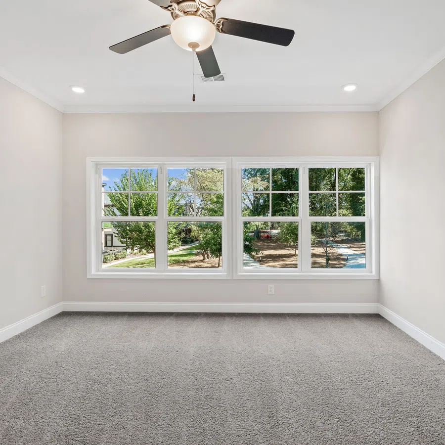 Empty room with gray carpet, large windows showing trees outside, white walls, and ceiling fan with light.
