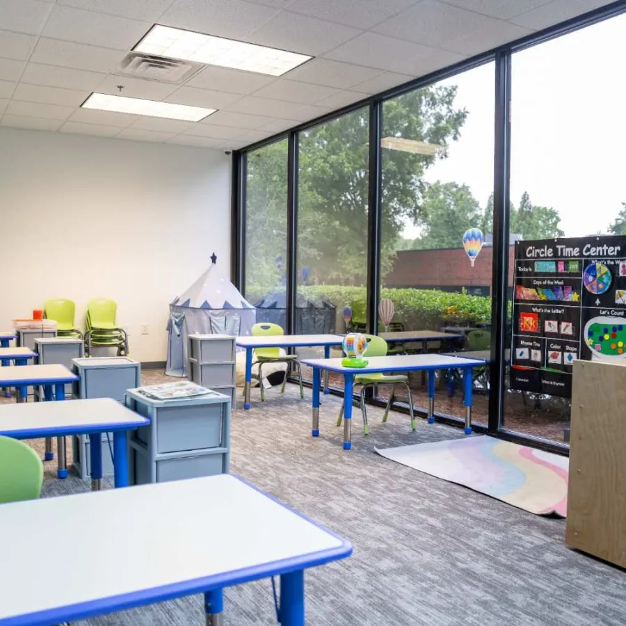 Bright and colorful early childhood classroom with small desks, green chairs, large windows, and educational toys.