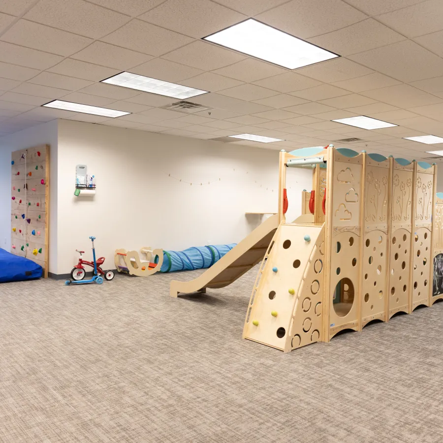 Bright indoor playroom with wooden climbing structure, slide, rock climbing wall, and toy vehicles on carpeted floor.