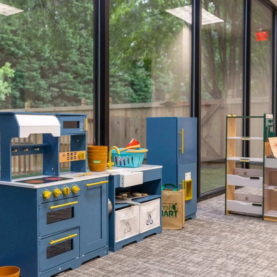 Children's play kitchen set with blue cabinets, toy food items, and play grocery shelves in a bright room.