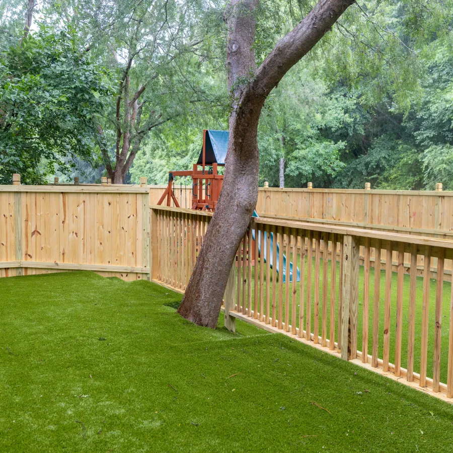 Backyard with green artificial grass, wooden fence, tree, playground slide, and trampoline under trees.