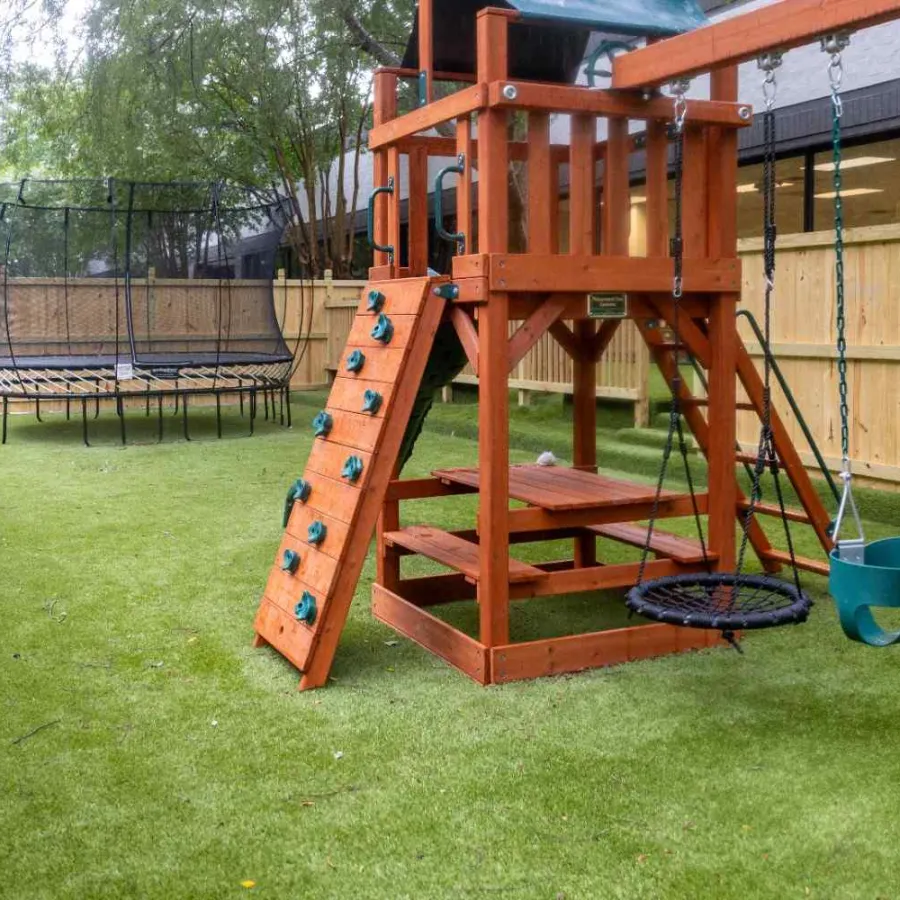 Backyard playground with wooden climbing structure, swings, and trampoline on green grass surrounded by a wooden fence.