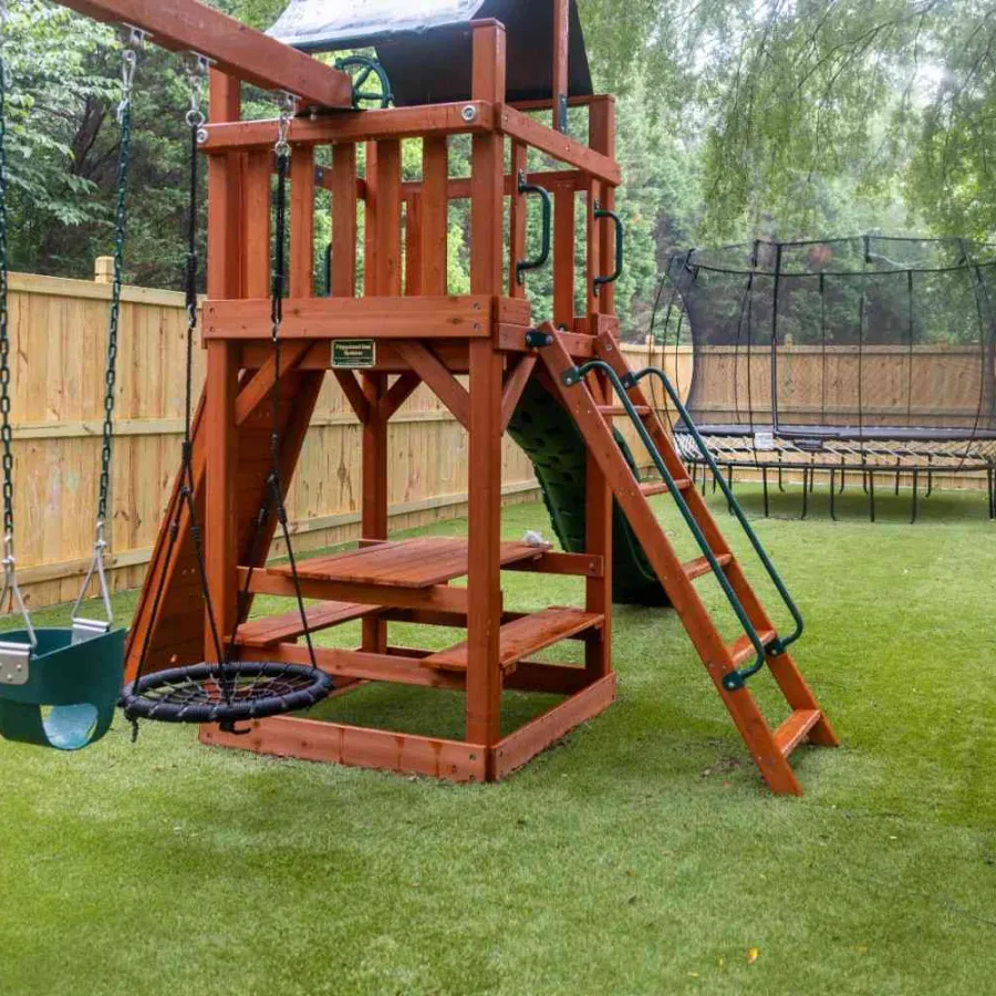 Children's wooden playground set with swings, slide, and trampoline on artificial grass in fenced backyard.