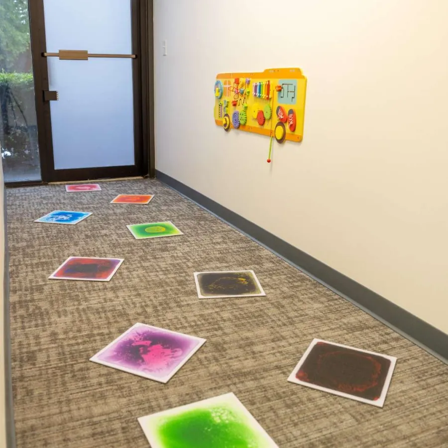 Indoor hallway with colorful square play mats on the floor and a yellow activity board on the wall near a frosted glass door.