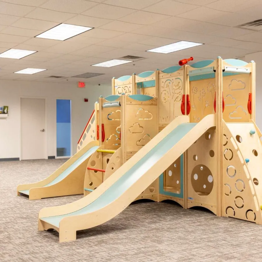 Indoor wooden playground with slides, climbing walls, and activity panels in a spacious room with carpeted floor.