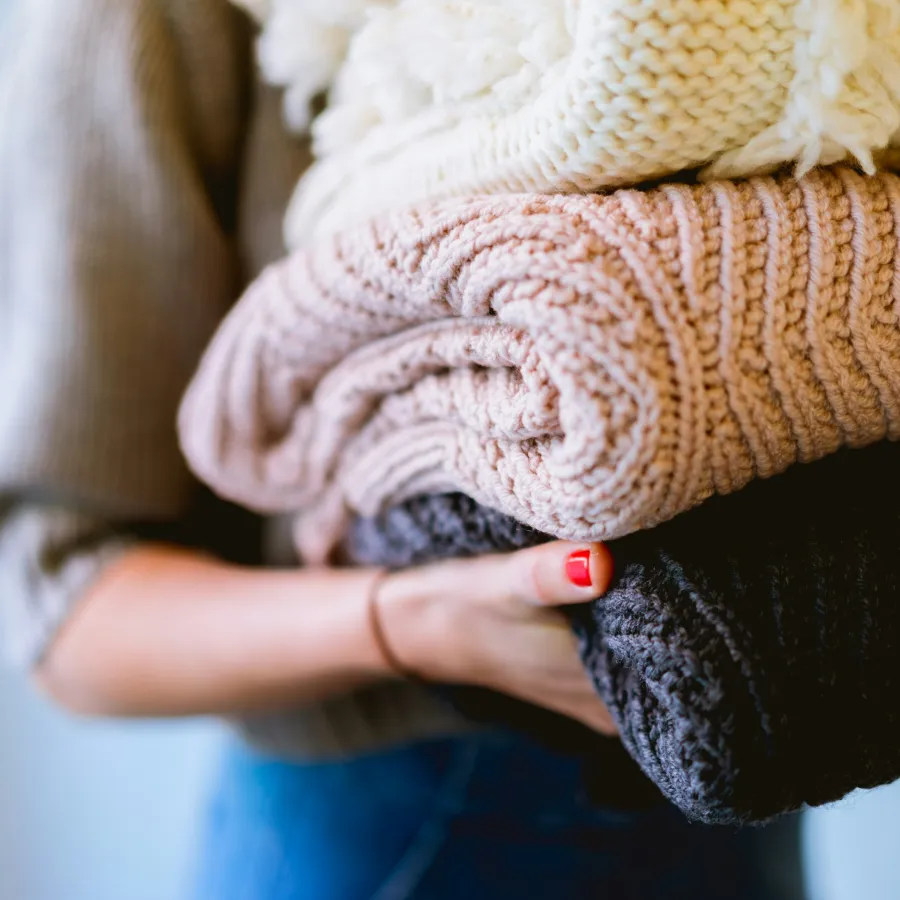 Person holding a stack of cozy folded knitted sweaters in cream, pink, and dark gray colors.