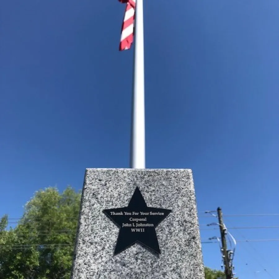 Granite memorial with a black star plaque honoring Corporal John L Johnson from WWII beneath a waving American flag.