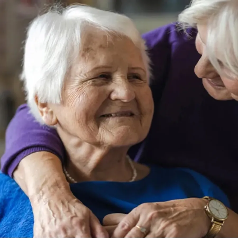 Elderly woman smiling warmly as another woman embraces her from behind showing care and affection.