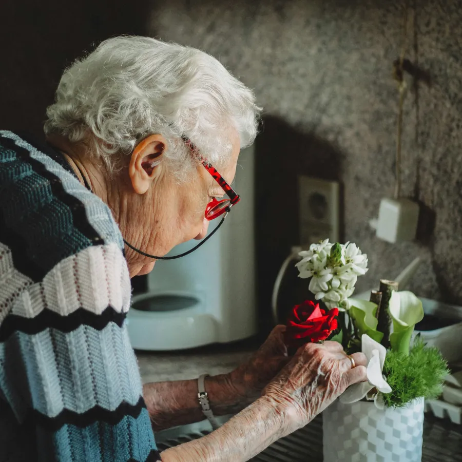 Elderly woman with white hair and red glasses arranging flowers in a white vase in a kitchen.