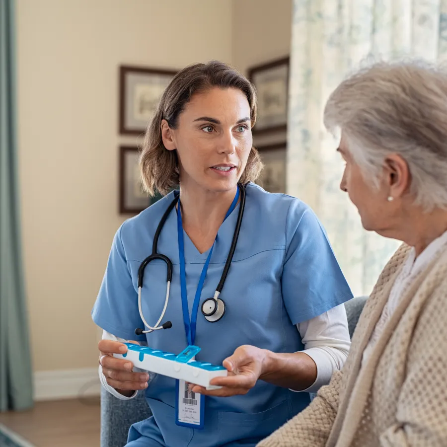 Nurse in blue scrubs explaining medication organizer to elderly woman in cozy living room setting
