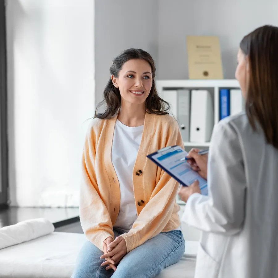 Female patient sitting on exam table smiling at doctor in white coat holding clipboard in bright medical office.