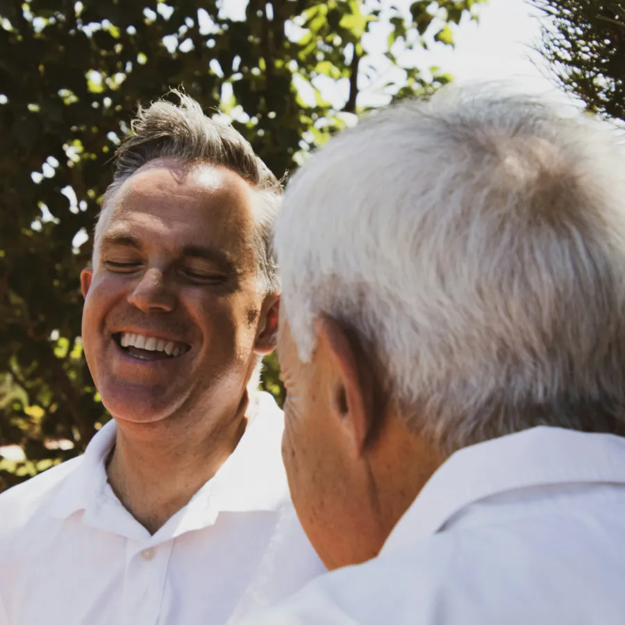 Two men in white shirts share a joyful conversation outdoors with greenery in the background.