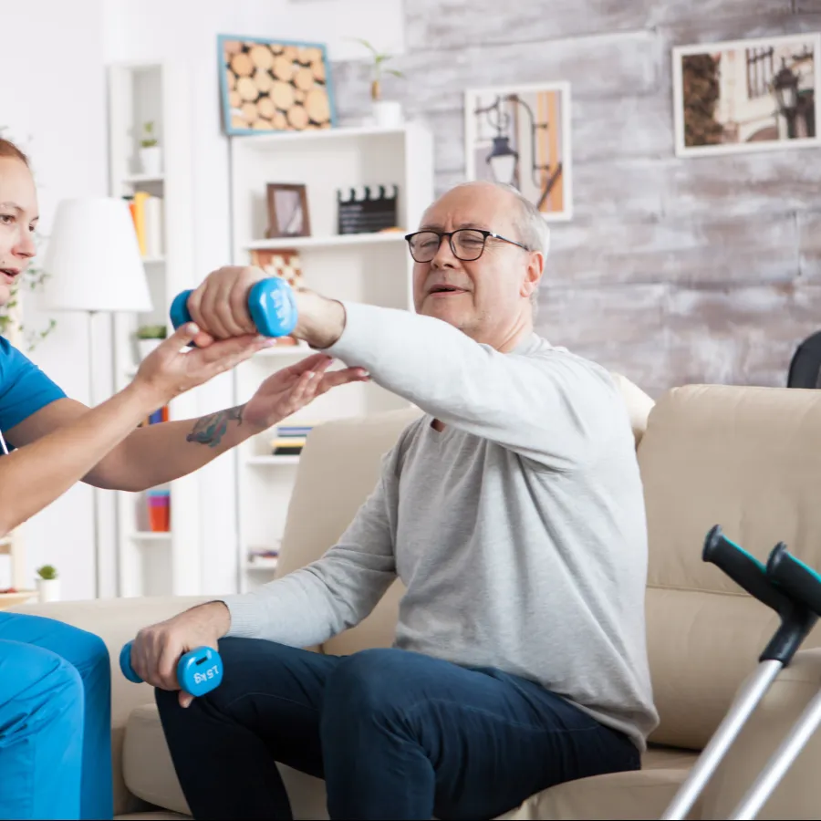 Healthcare worker assists elderly man with arm exercises using blue dumbbells in home rehabilitation.