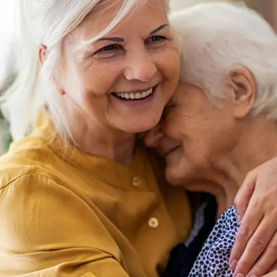 Smiling middle-aged woman warmly hugging elderly woman in a loving embrace indoors.