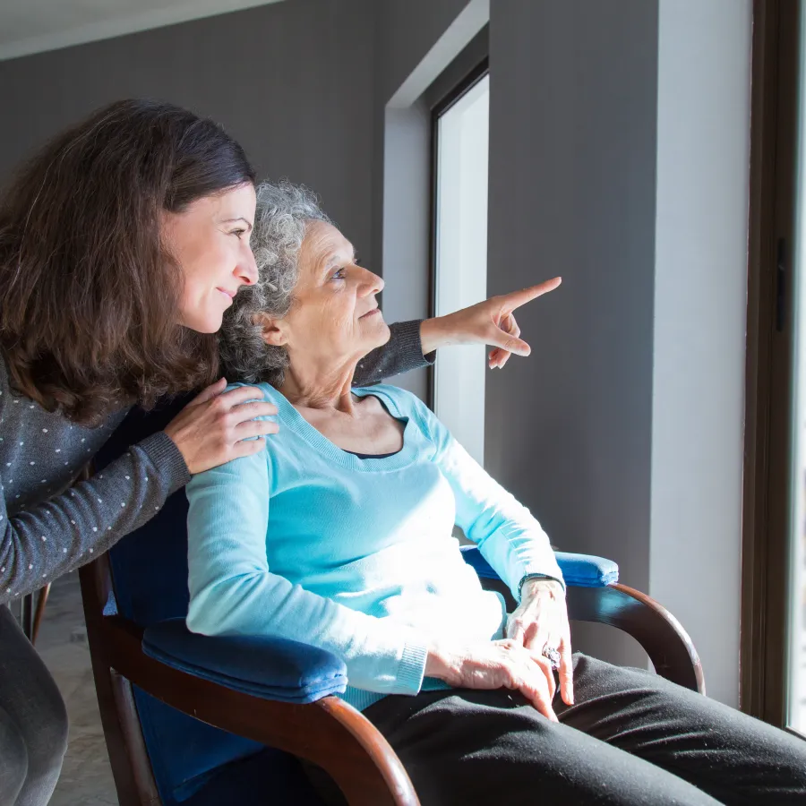 Elderly woman in wheelchair looking outside with younger woman gently supporting her, pointing out window view.