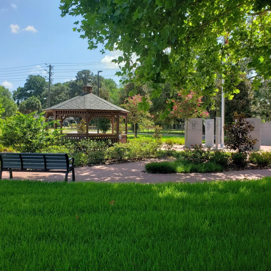Sunny park with green grass, a wooden gazebo, benches, and shaded trees under a blue sky with clouds.