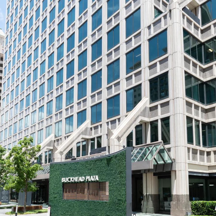 Modern glass office building with large windows and Buckhead Plaza sign with green hedge wall in foreground.