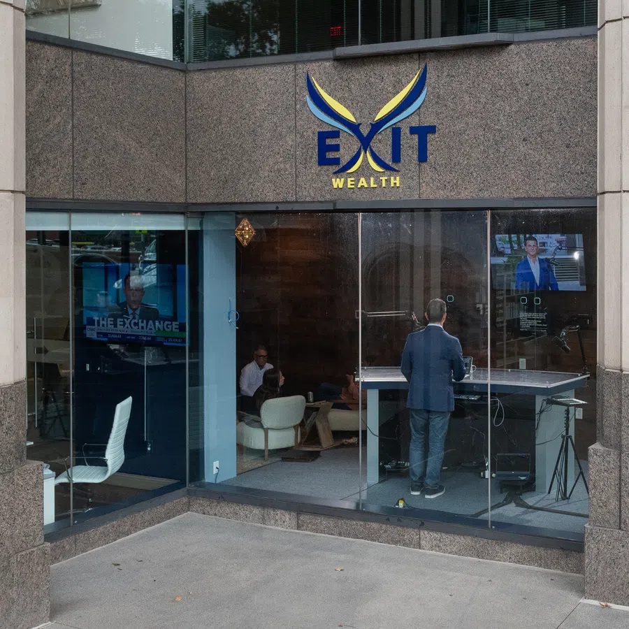 Exterior view of Exit Wealth office with glass walls showing people inside and a man standing at a desk with monitors.