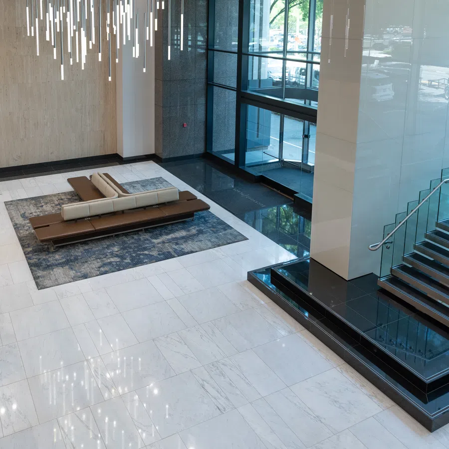 Modern building lobby with marble floor, glass stair railing, contemporary chandelier, and seating area on a rug.