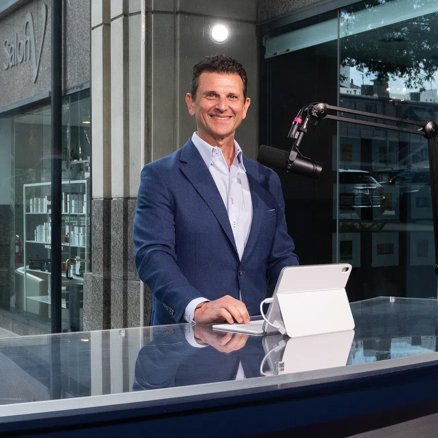 Smiling man in blue suit standing behind glass countertop with tablet and microphone setup outdoors.