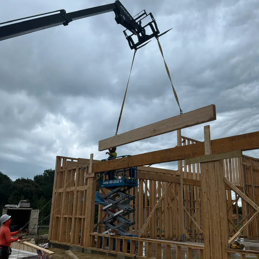 Crane lifting wooden beam during construction of wooden frame house under cloudy sky.
