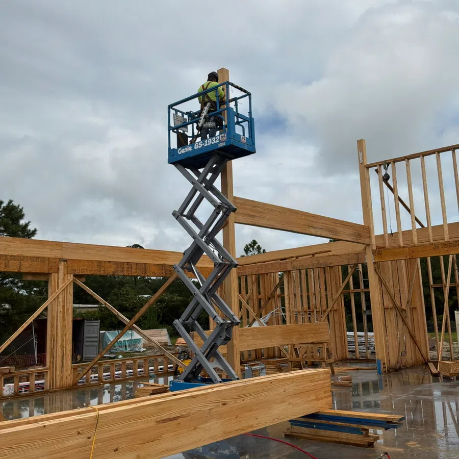 Construction worker on blue scissor lift installing wooden framing on a building under cloudy sky.