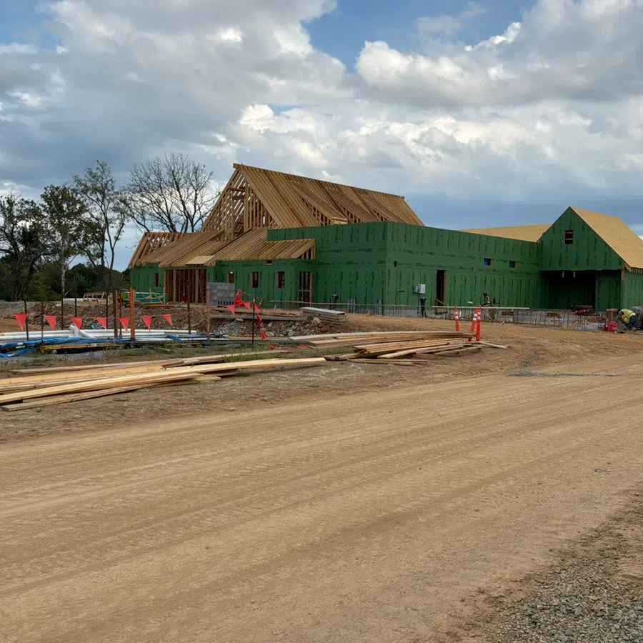 Large building under construction with wooden framework and green exterior panels on dirt site under cloudy sky.