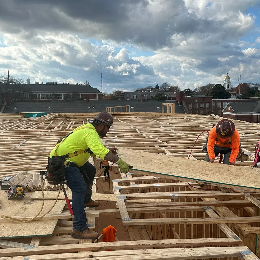 Construction workers install plywood sheathing on a wooden roof frame under a cloudy sky at a building site.
