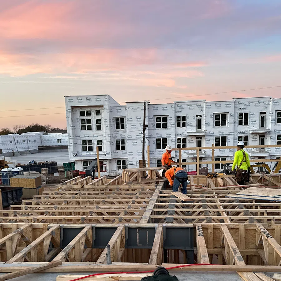 Construction workers building wooden framework of a residential development at sunset with white wrapped buildings in background.