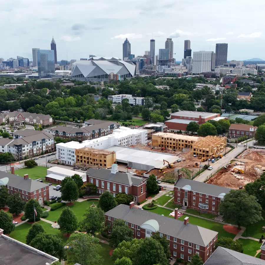 Aerial view of urban development with residential buildings, construction site, greenery, and a city skyline in the background.