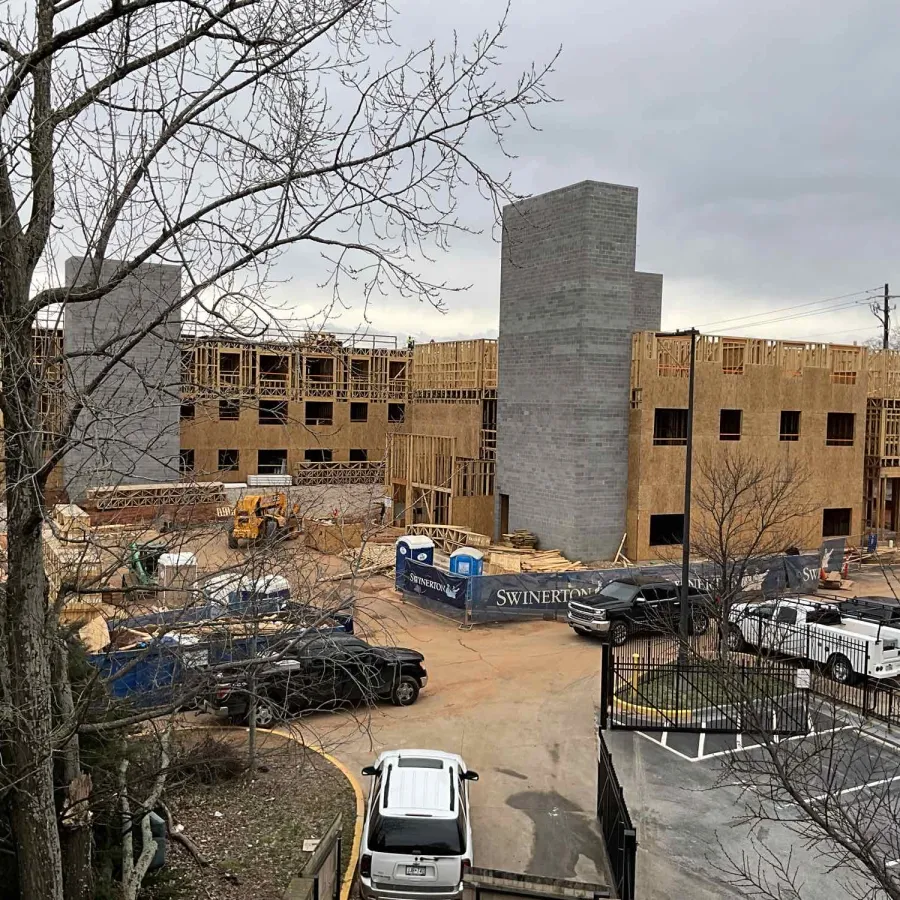 Multi-story building under construction with wooden framing, concrete structures, vehicles, and construction equipment on site.