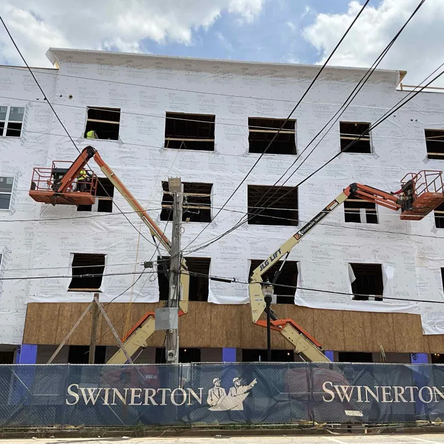 Construction site of a multi-story building with two cherry pickers and Swinerton fencing under a cloudy sky