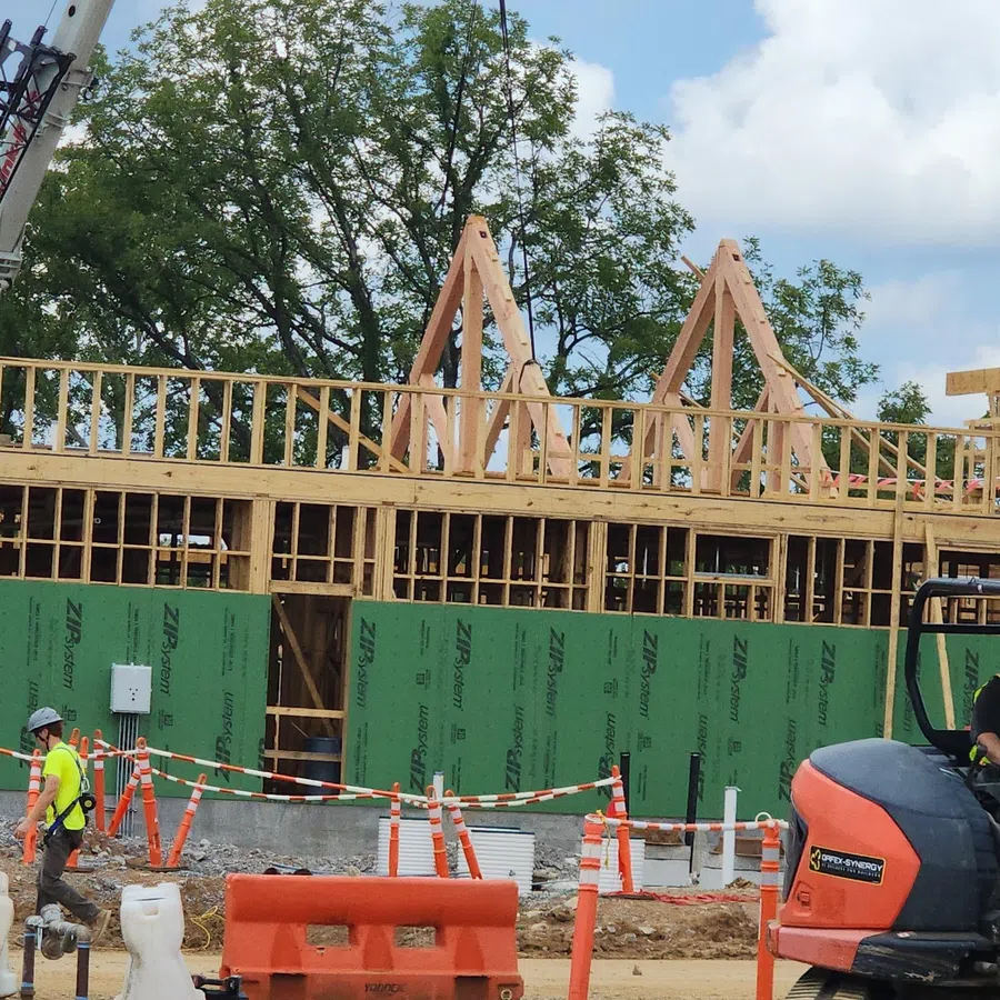 Construction site with wooden framing, workers in safety gear, and heavy machinery operating under a partly cloudy sky.