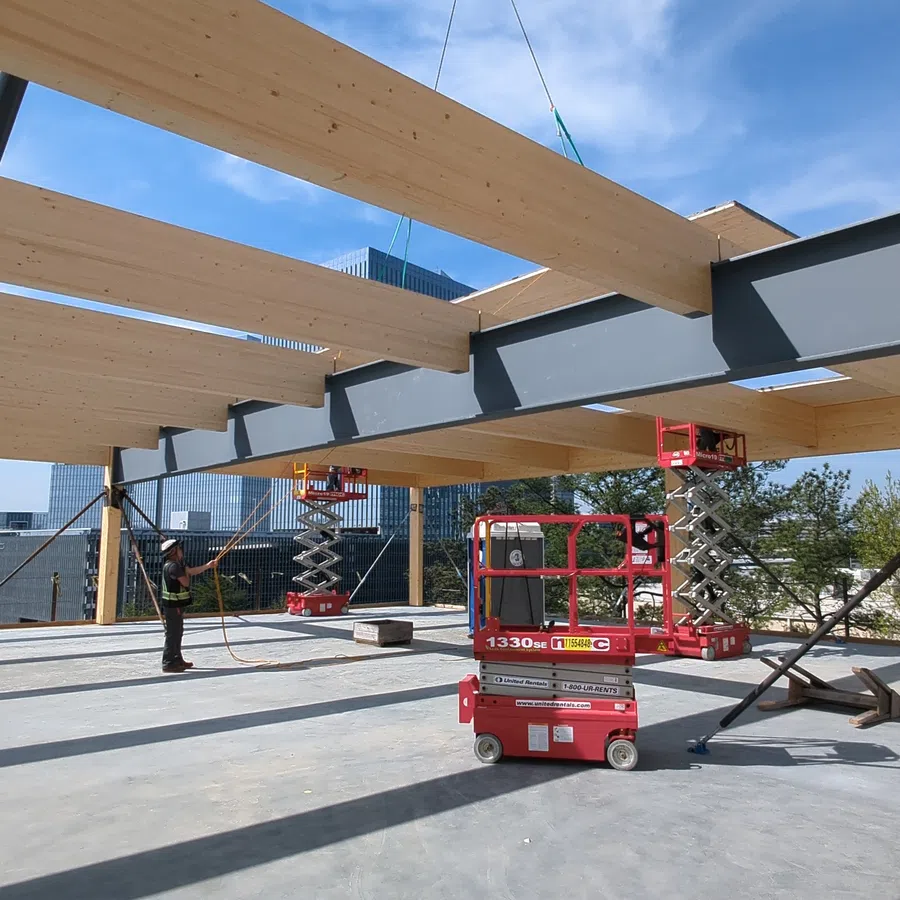 Construction site with wooden beams lifted by a crane and workers using red scissor lifts under blue sky.