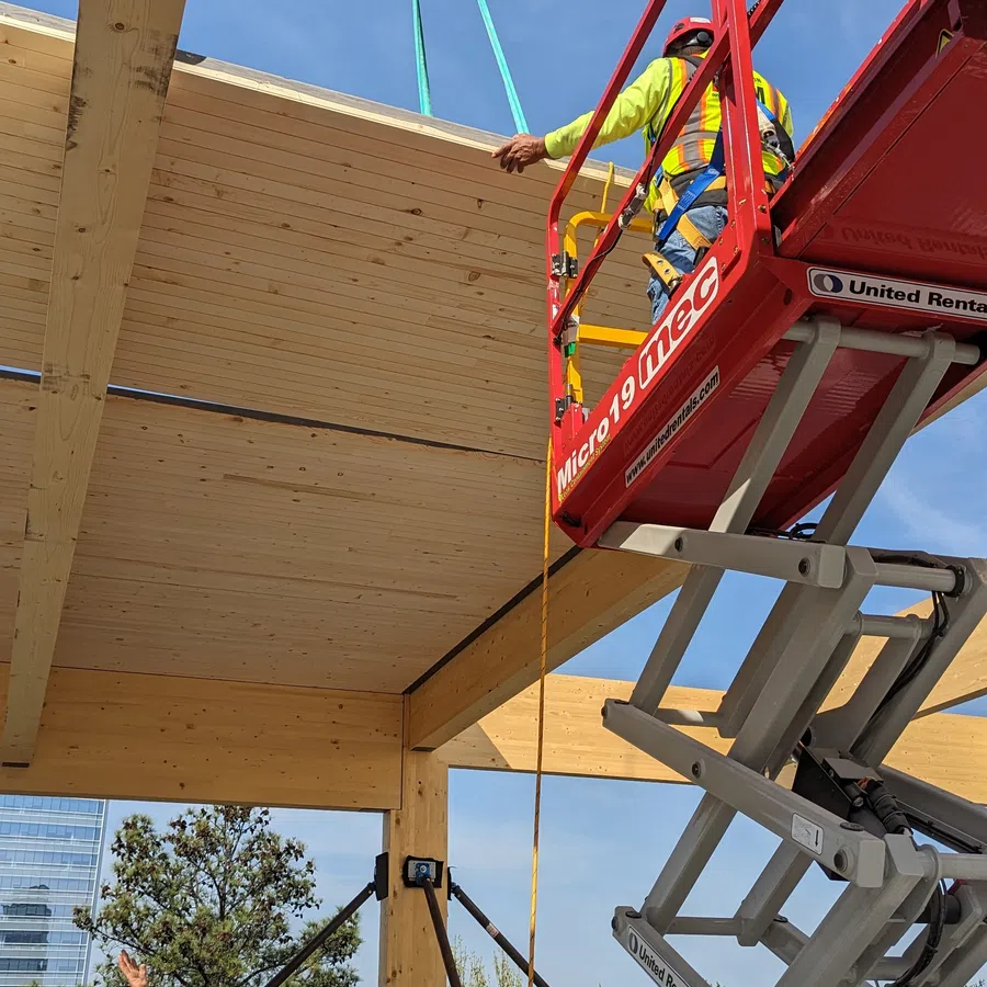 Construction workers installing large wooden beams using a red scissor lift on a sunny day.