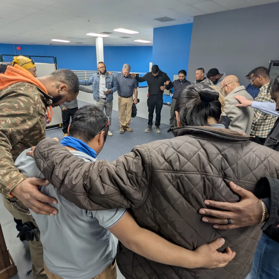 Group of diverse people standing in a circle with arms around each other in an indoor room.