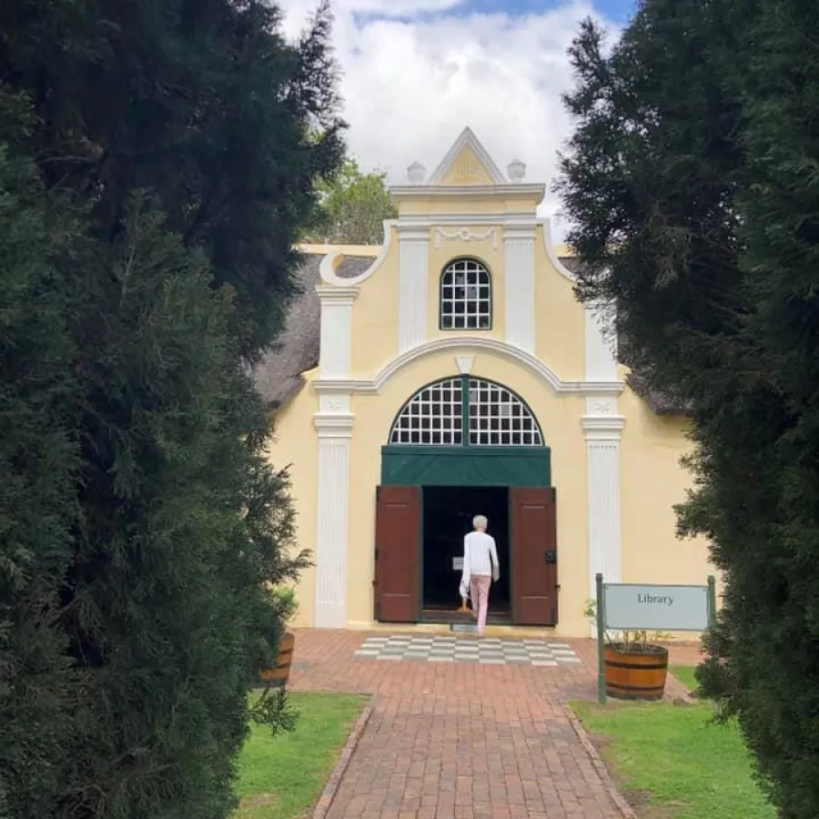 Person entering a historic yellow library building framed by tall trees on a brick pathway under blue sky.