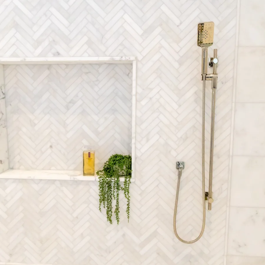 Modern shower setup with golden fixtures and a minimalist design featuring herringbone tiles and plants.