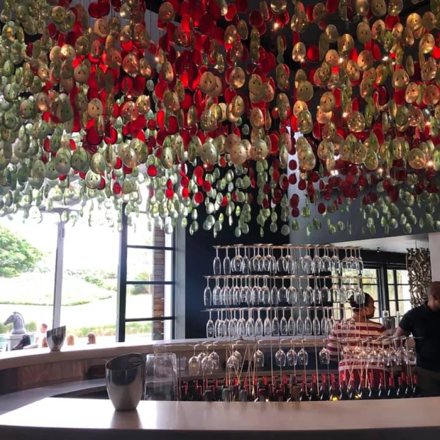 Modern bar interior with hanging glass art installation in red, green, and clear colors above counter and wine glasses.