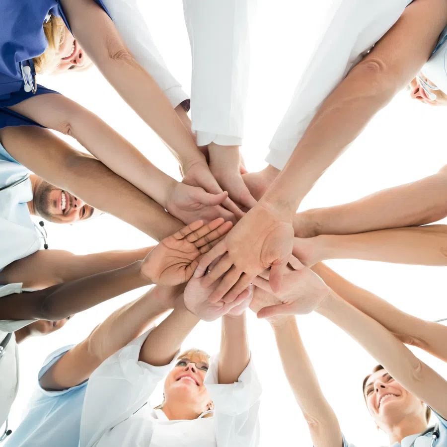 Diverse medical team in scrubs and lab coats stacking hands in teamwork gesture from below.