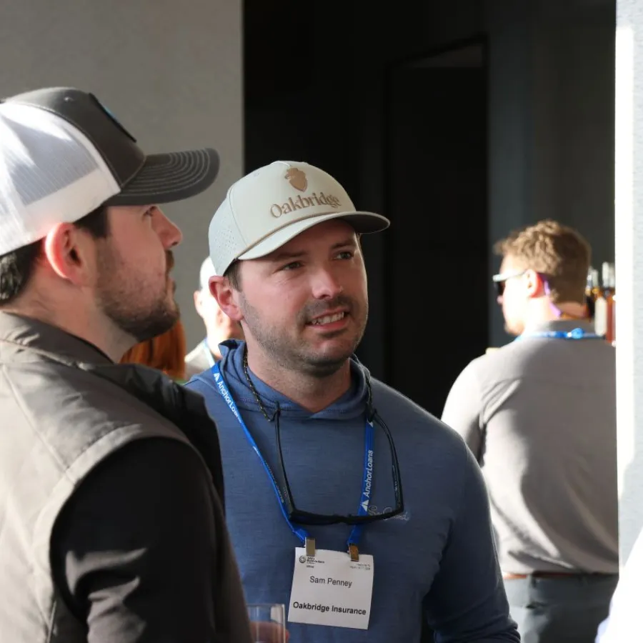 Two men wearing hats and name badges engaged in conversation at a professional event indoors.