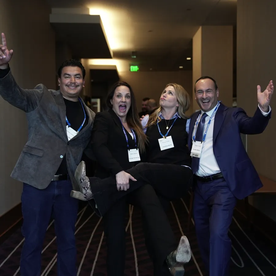 Four professionals at a conference hall posing playfully, smiling and wearing business attire with name badges.