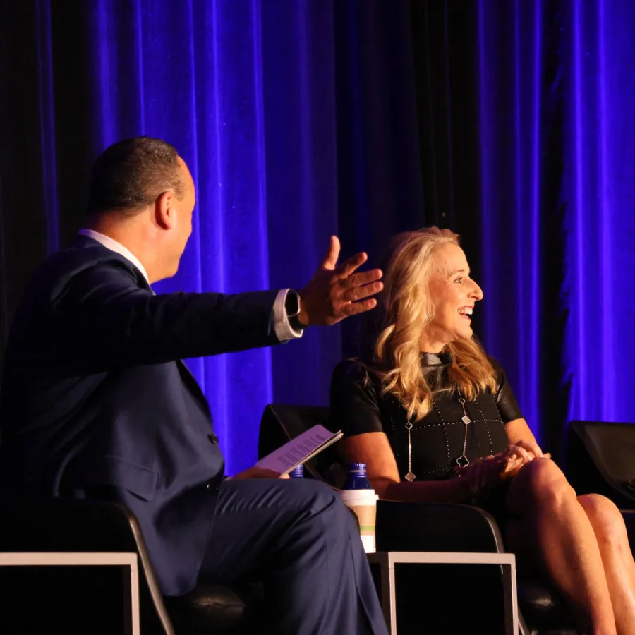 Two professionals engaged in lively discussion on stage with blue curtains in the background at a conference.