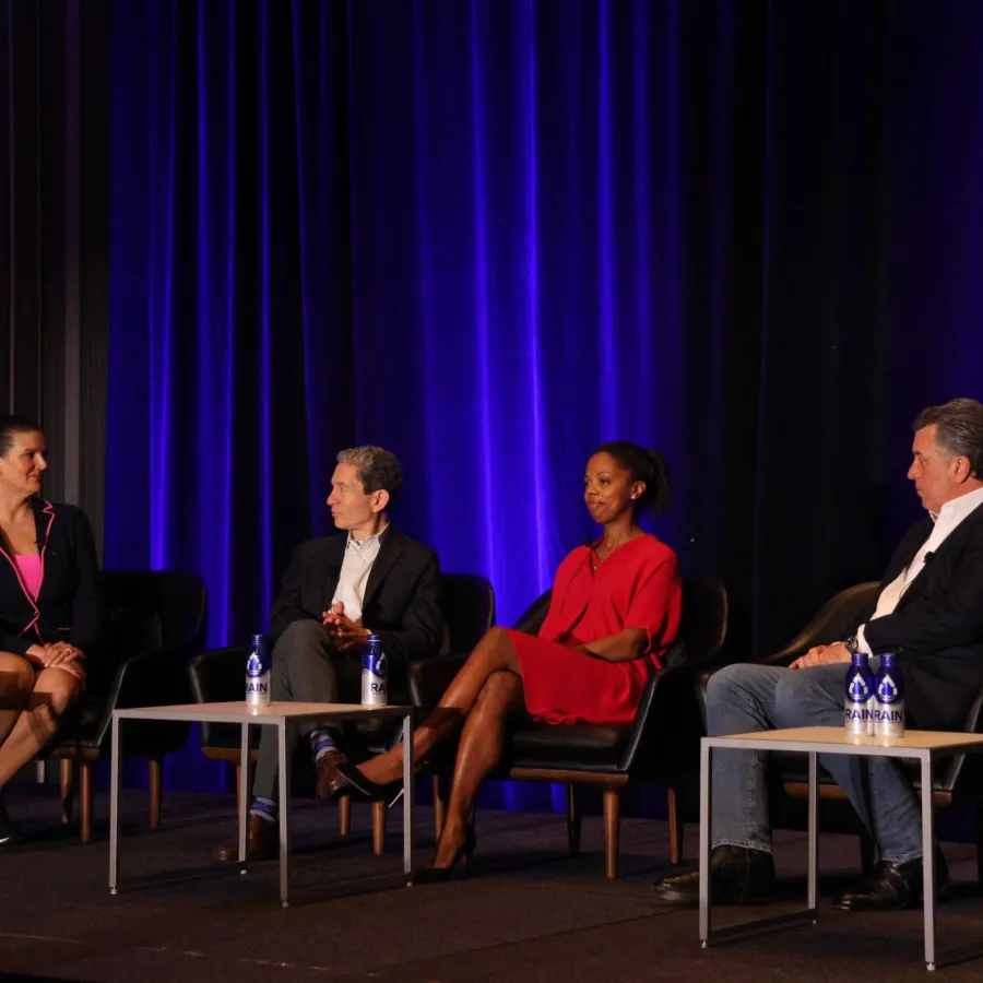 Four professionals seated on stage in a panel discussion with blue curtain backdrop and water bottles on tables.