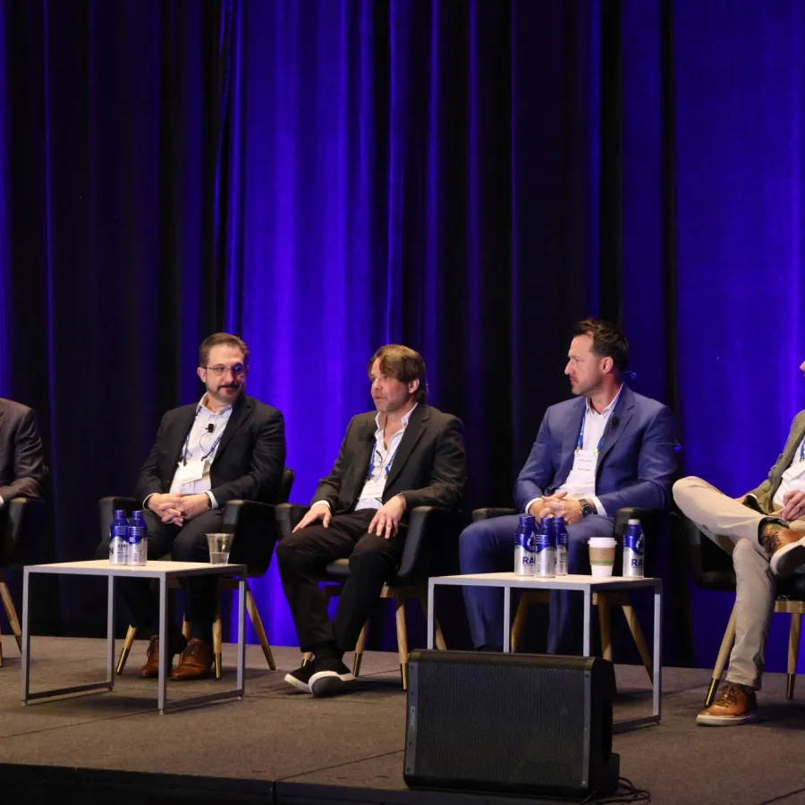 Five men in business attire seated on stage during a panel discussion with purple curtains in the background.