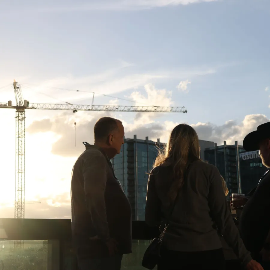Three people standing on a balcony at sunset with a city skyline and construction crane in the background.
