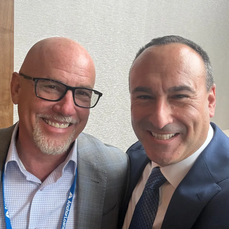 Two smiling men in business attire, one wearing glasses and an Anchor Loans badge, posing for a selfie indoors.